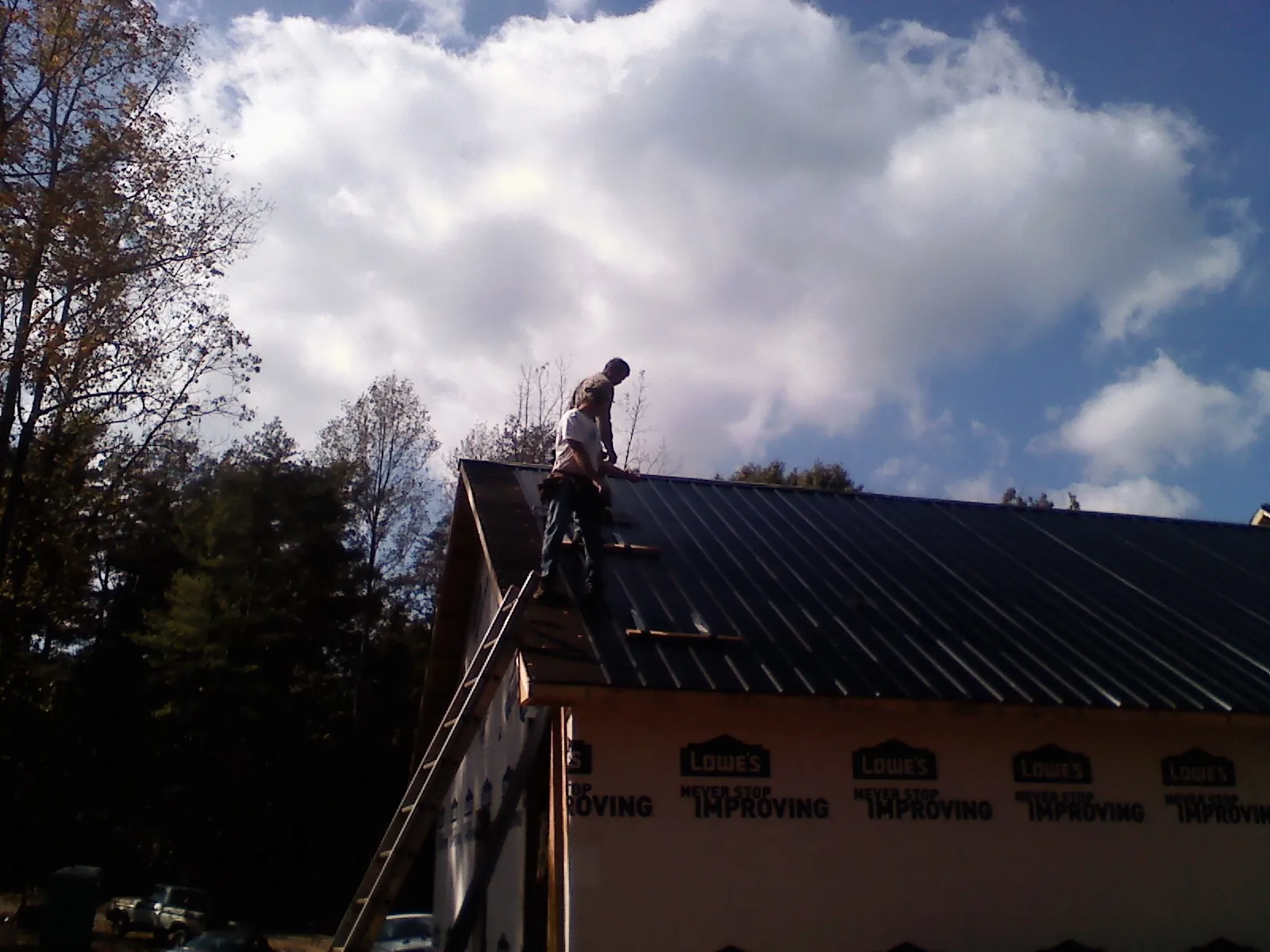 Roofer fastening dark metal panels high against a cloudy sky, from a Roof Inspection job in Lyons